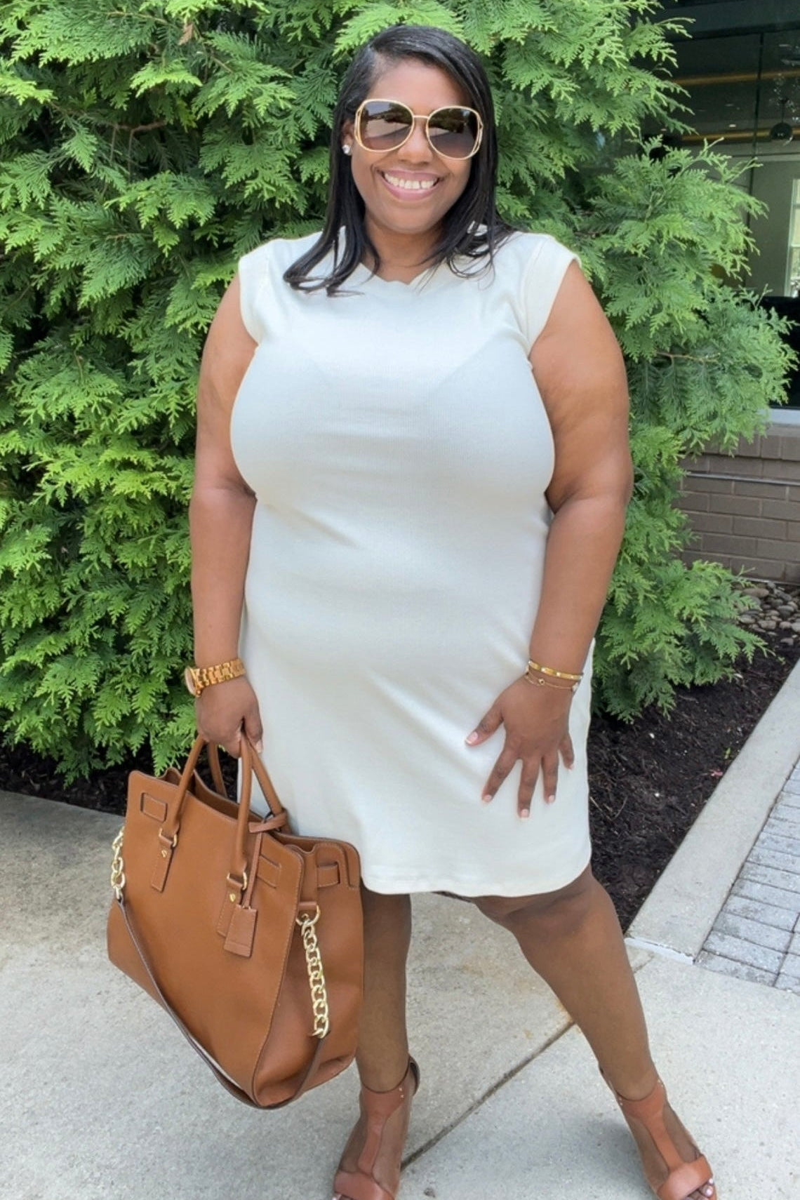 Woman in a white dress holding a brown handbag, standing outdoors with greenery in the background.