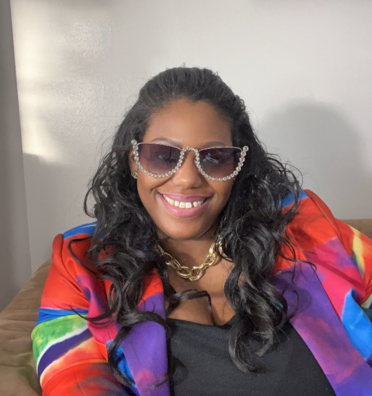 Woman smiling in rhinestone sunglasses and colorful blazer, sitting against a light wall.
