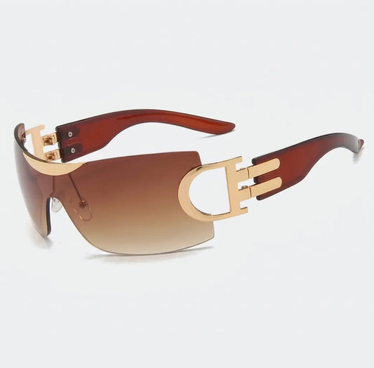 Rimless shield-style sunglasses with brown gradient lenses, gold side accents, and translucent brown arms displayed on a light gray background.