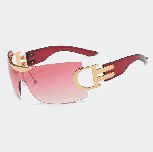 Rimless shield-style sunglasses with pink lenses, gold side accents, and translucent pink arms displayed on a light gray background.
