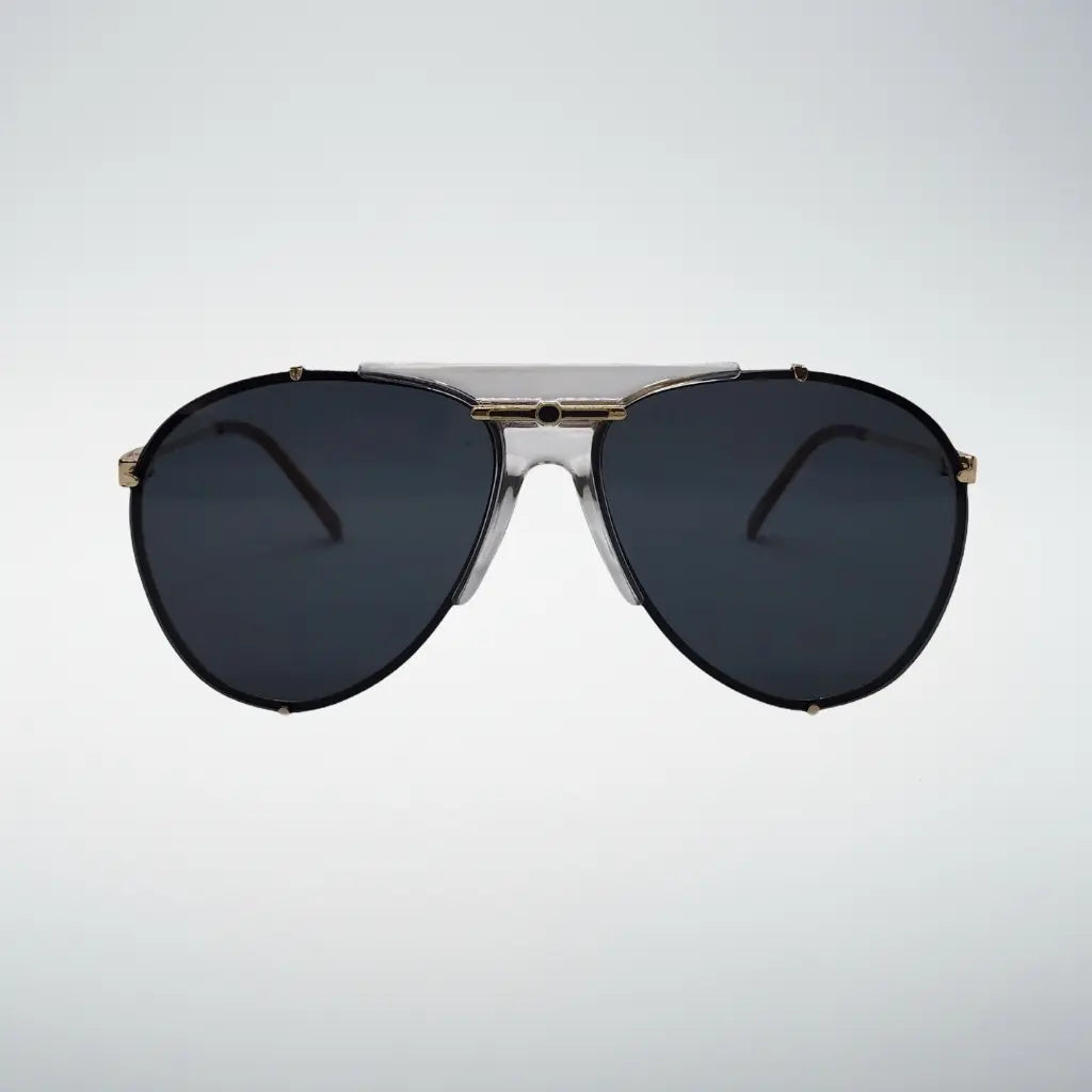 A pair of aviator-style sunglasses with dark gray lenses, a clear plastic upper brow bar, and gold metal accents on the frame, shown against a light gray gradient background.