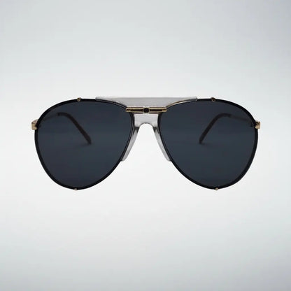 A pair of aviator-style sunglasses with dark gray lenses, a clear plastic upper brow bar, and gold metal accents on the frame, shown against a light gray gradient background.