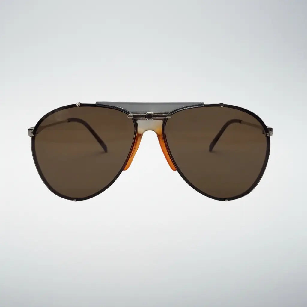A pair of aviator-style sunglasses with dark brown lenses, a silver-toned metal bridge, and orange acetate side accents, shown against a light gray gradient background.