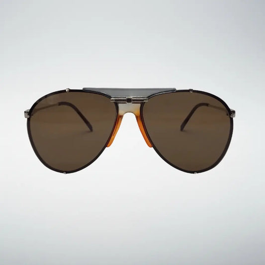 A pair of aviator-style sunglasses with dark brown lenses, a silver-toned metal bridge, and orange acetate side accents, shown against a light gray gradient background.