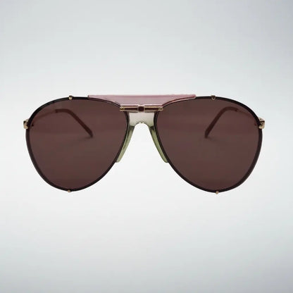A pair of aviator-style sunglasses with dark pink lenses, a metallic gold frame, and a dusty pink double bridge, shown against a light gray gradient background.